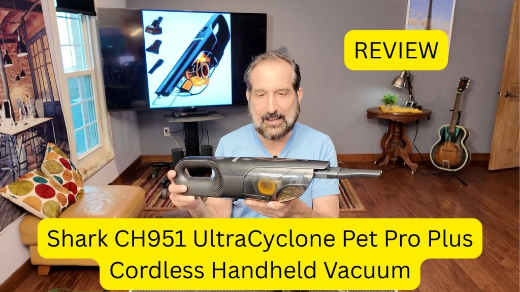 A man holding the Shark CH951 UltraCyclone Pet Pro Plus cordless handheld vacuum while reviewing it. A large screen displays vacuum attachments in the background.